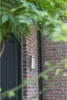 An outdoor wall light is mounted on a brick wall next to a dark door, with green foliage in the foreground. Produktbild-Vorschau