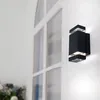 A black outdoor wall light is mounted on a white wooden wall next to a large white window. Voir les images du produit