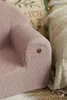 Close-up of a pink, fluffy children's armchair with a small wooden tag on the armrest. A beige blanket is visible in the background. Produktbild-Vorschau