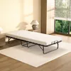 A foldable bed with a white mattress is placed on a light-colored rug in a room with a window. Voir les images du produit