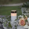A lit LED candle sits on a stone surface surrounded by ivy and roses. Produktbild-Vorschau