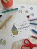 A collection of art supplies and handmade birthday cards on a wooden table. Voir les images du produit