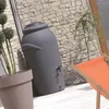 A dark gray rain barrel stands next to a stone wall on a patio, with a potted plant and an orange lounge chair visible. Produktbild-Vorschau