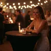 A woman in a white dress smiles at the camera while leaning on a table at an event with string lights in the background. Produktbild-Vorschau