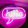 A neon sign shaped like a speech bubble with the word 'hello' inside, placed on a table next to a book and a perfume bottle. Anteprima dell’immagine prodotto