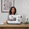 A woman is sitting at a wooden table with a sewing machine in front of her. Produktbild-Vorschau