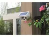 A white solar-powered outdoor camera is mounted on a textured brown wall, with green foliage and pink flowers growing around it. Produktbild-Vorschau