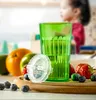 A green learning cup is placed on a wooden board with fruits and berries around it. A child is blurred in the background. Produktbild-Vorschau