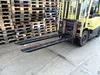 A yellow forklift with black fork extensions is parked in front of stacks of wooden pallets. Produktbild-Vorschau