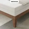 A close-up view of a mattress on a wooden bed frame, with a rug and concrete floor visible. Produktbild-Vorschau