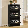 A black shoe cabinet with three open flip-down doors, displaying various shoes inside. The cabinet is placed against a cream-colored wall, with a pair of beige slippers on the floor in the foreground. A table and chair are visible in the background. Podgląd zdjęcia produktu