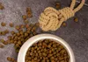 A bowl of dog food with kibble spilled around it and a rope toy on a textured gray surface. Produktbild-Vorschau