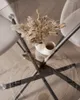 A close-up overhead view of a round glass-top dining table with a decorative vase of dried flowers on it. The table has a black metal base and is set on a light wood floor with a herringbone pattern. Two white chairs are partially visible at the edges of t Produktbild-Vorschau