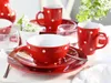 A set of red and white polka dot ceramic dishes, including mugs, bowls, and plates, arranged on a white tablecloth with a small bouquet of white flowers. Produktbild-Vorschau