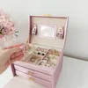 A pink jewelry box with several rings and earrings inside, with a hand reaching in to pick up a pair of earrings. Náhled obrázku produktu