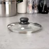 A glass pot lid with a black handle sits on a speckled gray countertop. Náhled obrázku produktu
