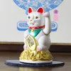 A white Maneki-neko (lucky cat) figurine is displayed on a dark surface, with a blurred blue and white patterned artwork in the background. Produktbild-Vorschau