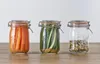 Three Kilner jars filled with preserved food items and spices are displayed on a wooden surface against a plain background. Anteprima dell’immagine prodotto