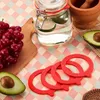A collection of red rubber sealing rings are arranged in the foreground, with a glass jar containing water and lemon slices in the midground. A bunch of red grapes on a wooden cutting board and a halved avocado are also visible. Anteprima dell’immagine prodotto