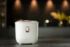 A white Lauben rice cooker with rose gold accents sits on a dark gray surface with a blurred background of warm lights and greenery. Podgląd zdjęcia produktu