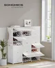 A white shoe rack with multiple compartments, some open and displaying shoes, is placed in a room with a window and a plant. Náhled obrázku produktu