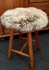 A round stool with a fluffy sheepskin cover sits on a dark carpet in front of a wooden cabinet. Produktbild-Vorschau