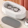 A white ultrasonic cleaner with glasses inside, placed on a table next to a gray mat and a watch. Produktbild-Vorschau
