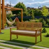 A wooden garden bench with a beige cushion sits on a lush green lawn in a backyard setting, with a hammock and pergola in the background. Produktbild-Vorschau