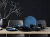A set of 16 Nordic Fjord Blue stoneware dishes, including plates and bowls, are arranged on a wooden table against a dark blue wall. A black vase with dry branches is placed to the left. Produktbild-Vorschau