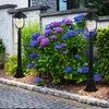 Two black outdoor lamps flank a large bush with blue and purple hydrangeas, set against a stone wall and a paved walkway. Produktbild-Vorschau