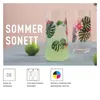 Two glasses with a tropical leaf and flower design, one filled with a light green beverage and ice, next to a lime on a blue tablecloth. Anteprima dell’immagine prodotto