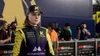 A female racing driver in a yellow and black racing suit and cap with a 'K' logo, standing in front of a crowd and cameras. Produktbild-Vorschau