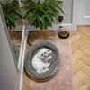 A small white dog is resting in its bed next to a dog bowl filled with food. A large potted plant is in the background. Produktbild-Vorschau