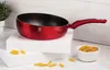 A red wok pan is displayed on white blocks, with penne pasta scattered around and in a small white bowl. Produktbild-Vorschau
