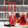 A pair of red and silver jar tongs are shown in the foreground, with two jars of red liquid in the midground. Several red apples are also visible in the background. Anteprima dell’immagine prodotto