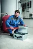 A man in a blue work uniform is kneeling next to an open silver toolbox, holding red pipe wrenches. Podgląd zdjęcia produktu