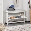 A white shoe bench with a cushioned top, featuring two flip-down shoe storage compartments. Hats are placed on top of the bench, and a basket with umbrellas is visible in the background. Produktbild-Vorschau