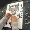 Hands assembling a wooden puzzle of an owl-like creature on a white surface, with scattered puzzle pieces around. Náhled obrázku produktu