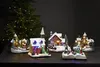 A collection of illuminated Christmas village houses and a church, set against a dark background with a blurred Christmas tree. Podgląd zdjęcia produktu