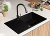 A modern black kitchen sink with a faucet, set on a white countertop with wooden cabinets below. Decorative elements like herbs and a mortar are placed on the countertop. Produktbild-Vorschau