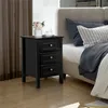 A black nightstand with three drawers sits next to a bed with gray bedding. Produktbild-Vorschau