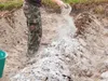 A person in camouflage pants is scattering white powder onto a pile of dirt and debris. Produktbild-Vorschau