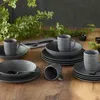A set of stoneware dishes and mugs arranged on a wooden table with greenery in the background. Náhľad obrázku produktu