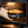 A hand wearing a beige oven mitt is placing a round cake pan with a cake inside into a lit oven. Anteprima dell’immagine prodotto