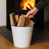 A hand is holding a white metal bucket filled with firewood next to a fireplace. Voir les images du produit