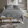 A neatly made bed with grey striped bedding, a wooden stool with books, and a sheepskin rug on the floor, in a minimalist bedroom setting. Produktbild-Vorschau