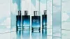 Four bottles of perfume are displayed on a reflective surface with a background of mirrored panels showing a cloudy sky. Voir les images du produit