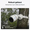 A white security camera is mounted on a tree trunk during a rain shower. Produktbild-Vorschau