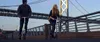 Two people riding scooters on a bridge with a large bridge structure in the background. Produktbild-Vorschau