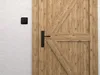 A black door handle and a black light switch are shown on a white wall next to a wooden door. Produktbild-Vorschau